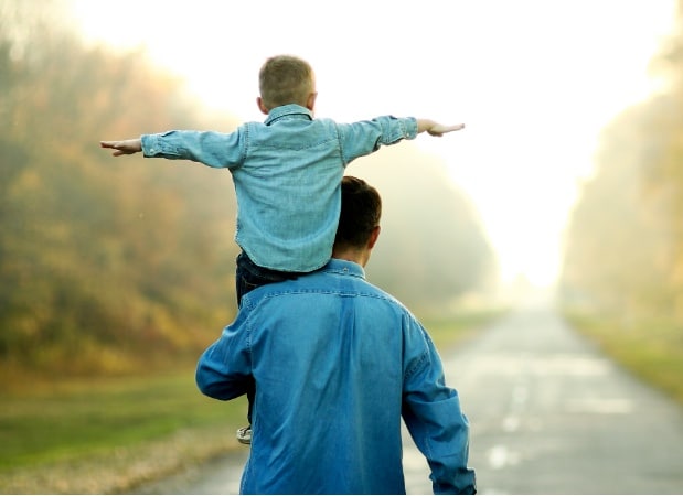 a boy with arms outstretched riding on the shoulders of his father